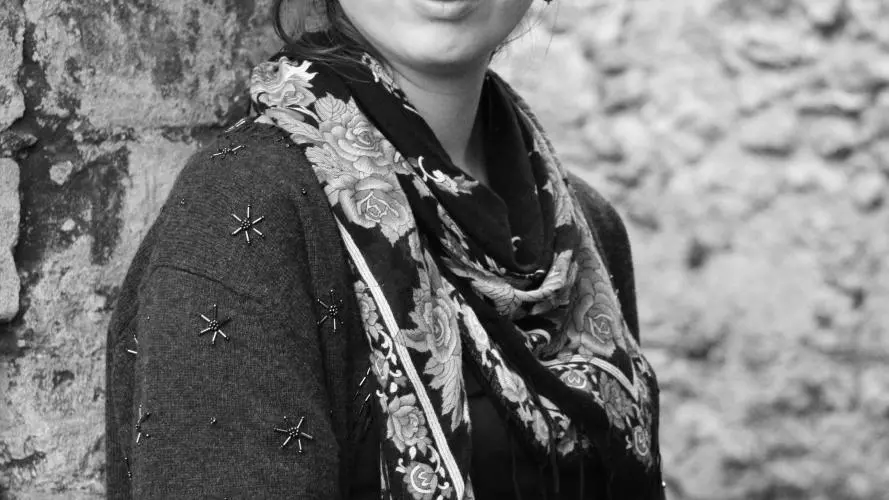 A black & white photograph of Penny Boxall, who stands with her back against a tree trunk. Her face is turned directly towards the viewer, and she is dressed in dark clothing with a bright floral scarf and drop earrings.