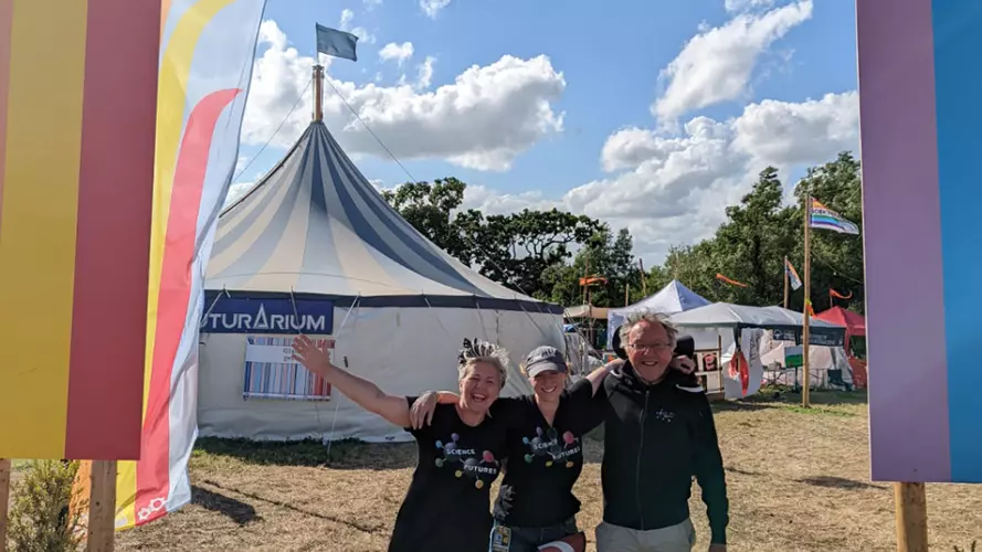 Three members of the Science Futures team stand beneath the rainbow-coloured wooden entrance to the Science Futures Area, on a sunny day at Glastonbury 2022. The team are smiling and waving.